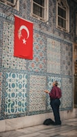 A man stands facing a wall adorned with intricate blue and white tiles featuring various patterns. Above him, a red flag with a white star and crescent hangs. The man appears to be engaged in prayer or meditation.