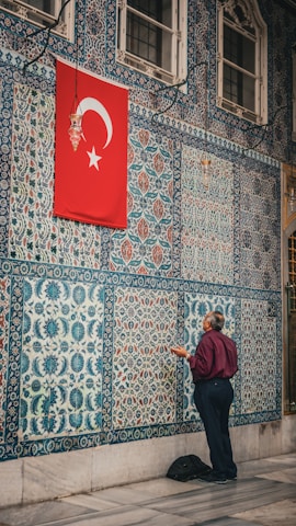 A man stands facing a wall adorned with intricate blue and white tiles featuring various patterns. Above him, a red flag with a white star and crescent hangs. The man appears to be engaged in prayer or meditation.