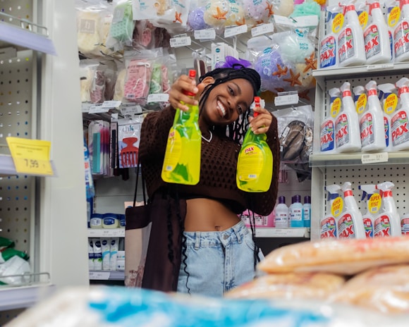 A person is smiling and holding two bottles of cleaning product in a store aisle. Behind them are various household items like detergents, hygiene products, and bath sponges displayed on shelves. The atmosphere appears cheerful and casual.