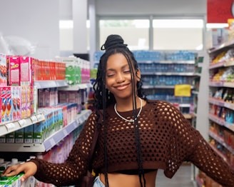 A person with long braided hair and a netted brown sweater stands smiling in a grocery store aisle. The shelves are stocked with colorful boxes and cartons, suggesting a variety of products.
