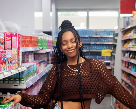 A person with long braided hair and a netted brown sweater stands smiling in a grocery store aisle. The shelves are stocked with colorful boxes and cartons, suggesting a variety of products.