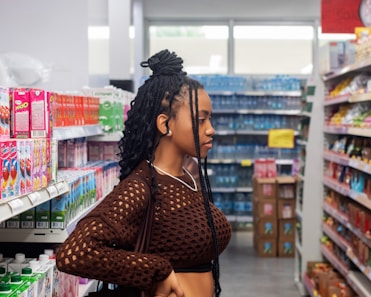 A person with braided hair stands in a grocery store aisle, browsing products. The shelves are stocked with colorful boxes, bottles, and snacks. The store is brightly lit, with a variety of items visible in the background.