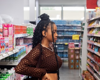 A person with braided hair stands in a grocery store aisle, browsing products. The shelves are stocked with colorful boxes, bottles, and snacks. The store is brightly lit, with a variety of items visible in the background.