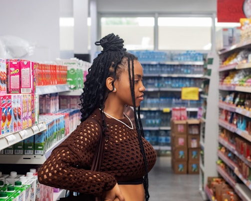 A person with braided hair stands in a grocery store aisle, browsing products. The shelves are stocked with colorful boxes, bottles, and snacks. The store is brightly lit, with a variety of items visible in the background.