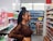 A person with braided hair stands in a grocery store aisle, browsing products. The shelves are stocked with colorful boxes, bottles, and snacks. The store is brightly lit, with a variety of items visible in the background.