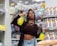 A person stands in a store aisle holding two cleaning spray bottles, surrounded by various household products. The shelves are stocked with bath sponges, personal care items, and a selection of cleaning products. Price tags are visible on the shelves.