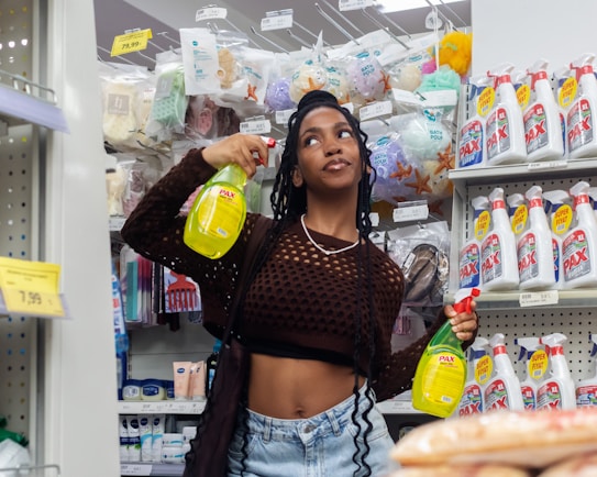 A person stands in a store aisle holding two cleaning spray bottles, surrounded by various household products. The shelves are stocked with bath sponges, personal care items, and a selection of cleaning products. Price tags are visible on the shelves.