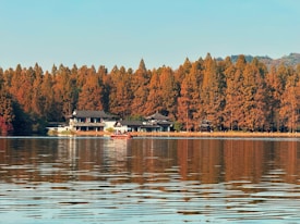 A tranquil scene of a lakeside with traditional-style buildings partially hidden among tall, orange-colored trees. The clear water of the lake reflects the autumn foliage and the sky. A small boat with people is seen near the shore, adding a peaceful human element to the natural setting.