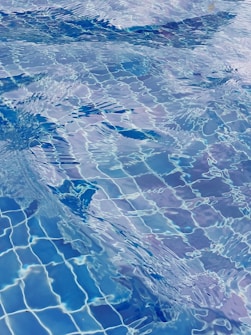 The image features the surface of a swimming pool with shimmering water ripples. The water creates a dynamic pattern with visible reflections, showcasing various shades of blue and hints of white. The vividness of the scene evokes a sense of movement and fluidity.