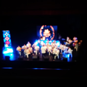 A blurred image of a stage with multiple people, possibly a choir or an orchestra, surrounded by bright blue lights. A central circular decorative element is visible in the background.