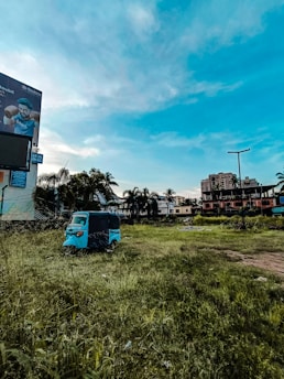 An open grassy field with a blue auto rickshaw parked near the center. In the background, there is a billboard featuring a person, some palm trees, and various buildings including a tall structure with a flat roof. The sky is predominantly clear with a few clouds and a vibrant blue hue.