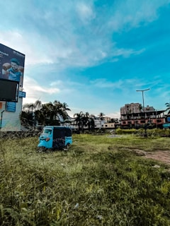 An open grassy field with a blue auto rickshaw parked near the center. In the background, there is a billboard featuring a person, some palm trees, and various buildings including a tall structure with a flat roof. The sky is predominantly clear with a few clouds and a vibrant blue hue.