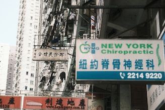 A busy urban street with high-rise residential buildings in the background. Various signs are displayed prominently, including one for 'New York Chiropractic' in both English and Chinese. Another sign is in Chinese characters, and there are several balconies with hanging laundry.