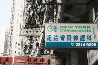 A busy urban street with high-rise residential buildings in the background. Various signs are displayed prominently, including one for 'New York Chiropractic' in both English and Chinese. Another sign is in Chinese characters, and there are several balconies with hanging laundry.