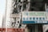 A busy urban street with high-rise residential buildings in the background. Various signs are displayed prominently, including one for 'New York Chiropractic' in both English and Chinese. Another sign is in Chinese characters, and there are several balconies with hanging laundry.