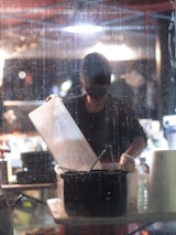 A person stands behind a wet glass, working with kitchen utensils under a bright overhead light. A large translucent container is held above a black pot, while various items like a roll of paper towels and a water bottle are on the table. The background appears busy and blurred with lights and shapes.