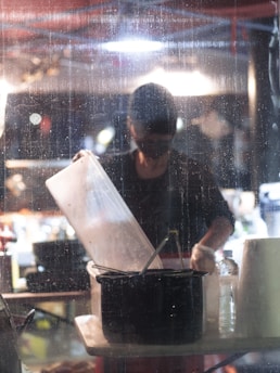 A person stands behind a wet glass, working with kitchen utensils under a bright overhead light. A large translucent container is held above a black pot, while various items like a roll of paper towels and a water bottle are on the table. The background appears busy and blurred with lights and shapes.