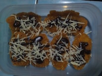 A plastic container holds several pieces of a fried golden-brown snack topped with chocolate syrup and shredded cheese.