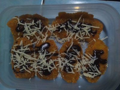 A plastic container holds several pieces of a fried golden-brown snack topped with chocolate syrup and shredded cheese.