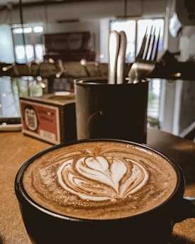 A cup of coffee with intricate latte art sits on a wooden table. In the background, there is a container holding utensils and a menu stand, with a blurred modern cafe interior.