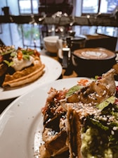 A close-up view of two plates of gourmet food, likely brunch items, garnished with fresh vegetables and sauces. One of the plates has waffles topped with whipped cream and fresh greens. In the background, there are several cups, including one with a frothy latte. The setting appears to be within a cozy café with natural light coming through the windows.