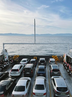 A ferry is carrying several cars parked in neat rows, crossing a calm, expansive body of water under a bright blue sky. On the sides, there are motorcycles lined up. Distant hills are visible on the horizon.