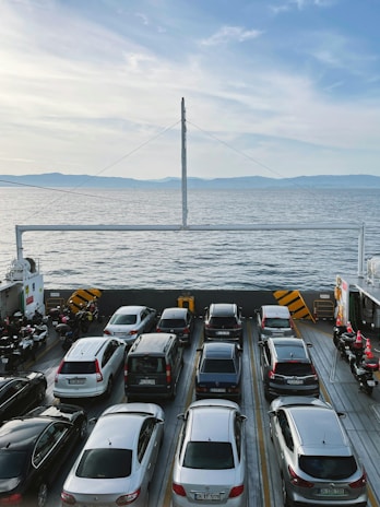 A ferry is carrying several cars parked in neat rows, crossing a calm, expansive body of water under a bright blue sky. On the sides, there are motorcycles lined up. Distant hills are visible on the horizon.