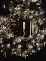 A decorative wreath made of evergreen branches adorned with small white lights. It is illuminated in a dark setting, creating a warm and festive atmosphere. In the background, a portion of what appears to be a doorway or window with soft lighting is visible.