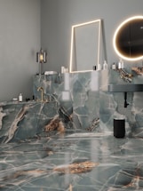 A modern bathroom features luxurious green and white marble surfaces throughout, including the walls, countertop, and floor. There is a rectangular mirror with a soft glow of LED lighting surrounding it and a circular mirror emits a warm ambient light. The gold fixtures, including a faucet, add an elegant touch. Various bottles and toiletries are neatly arranged on the countertop. The overall design is sleek and sophisticated.