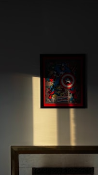 A framed comic-style illustration featuring a superhero with a shield, prominently displayed on a wall. A shaft of light illuminates the image, casting shadows and highlighting the superhero's distinctive colors. The artwork is positioned above a fireplace with a light and dark contrasting effect due to the lighting.