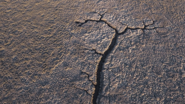 Aerial view of a barren, cracked landscape with a prominent fissure running through the center. The ground surface appears arid and textured, with irregular patterns formed by the dried earth.