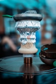 A close-up of a hookah setup, featuring a bowl wrapped with aluminum foil and coals on top in a metal screen. The hookah base is placed on a circular metallic surface, and there are blurred elements in the background, suggesting an indoor or café environment.