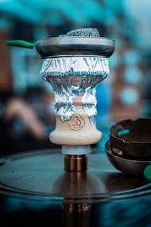 A close-up of a hookah setup, featuring a bowl wrapped with aluminum foil and coals on top in a metal screen. The hookah base is placed on a circular metallic surface, and there are blurred elements in the background, suggesting an indoor or café environment.
