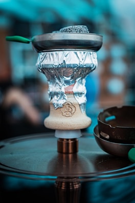A close-up of a hookah setup, featuring a bowl wrapped with aluminum foil and coals on top in a metal screen. The hookah base is placed on a circular metallic surface, and there are blurred elements in the background, suggesting an indoor or café environment.