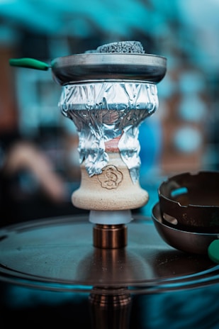 A close-up of a hookah setup, featuring a bowl wrapped with aluminum foil and coals on top in a metal screen. The hookah base is placed on a circular metallic surface, and there are blurred elements in the background, suggesting an indoor or café environment.