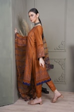 A person is wearing a traditional brown outfit with intricate patterns and a long embellished scarf. The outfit is complemented by neutral sandals and subtle jewelry. The background features a greenish wall with decorative molding and dried pampas grass.