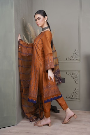 A person is wearing a traditional brown outfit with intricate patterns and a long embellished scarf. The outfit is complemented by neutral sandals and subtle jewelry. The background features a greenish wall with decorative molding and dried pampas grass.