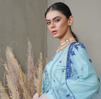 A person stands confidently in traditional attire, featuring intricate blue embroidery on a light blue fabric. The outfit consists of a long-sleeved top and a matching dupatta draped over the shoulder. The person wears elegant jewelry, including earrings and layered necklaces. Behind them, there are decorative pampas grass arrangements against a neutral wall.