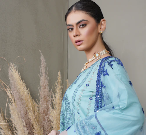 A person stands confidently in traditional attire, featuring intricate blue embroidery on a light blue fabric. The outfit consists of a long-sleeved top and a matching dupatta draped over the shoulder. The person wears elegant jewelry, including earrings and layered necklaces. Behind them, there are decorative pampas grass arrangements against a neutral wall.