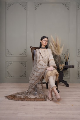 A woman is sitting elegantly on a wooden chair with intricate carvings. She is wearing a traditional embroidered outfit with a sheer dupatta. The setting includes a decorative backdrop with wall paneling featuring corner ornaments. Beside her is a small table with a vase containing pampas grass and decorative sticks.
