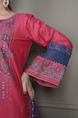 A detailed close-up of a garment with intricate embroidery. The fabric is a vibrant pink with elaborate patterns and embellishments highlighting the design. The sleeve features decorative blue lace with floral motifs, and the person is wearing large hoop earrings.