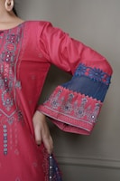 A detailed close-up of a garment with intricate embroidery. The fabric is a vibrant pink with elaborate patterns and embellishments highlighting the design. The sleeve features decorative blue lace with floral motifs, and the person is wearing large hoop earrings.