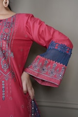 A detailed close-up of a garment with intricate embroidery. The fabric is a vibrant pink with elaborate patterns and embellishments highlighting the design. The sleeve features decorative blue lace with floral motifs, and the person is wearing large hoop earrings.
