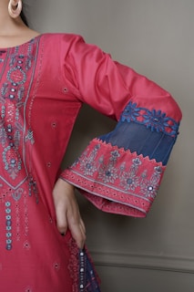A detailed close-up of a garment with intricate embroidery. The fabric is a vibrant pink with elaborate patterns and embellishments highlighting the design. The sleeve features decorative blue lace with floral motifs, and the person is wearing large hoop earrings.