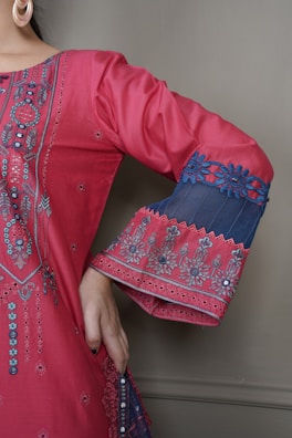 A detailed close-up of a garment with intricate embroidery. The fabric is a vibrant pink with elaborate patterns and embellishments highlighting the design. The sleeve features decorative blue lace with floral motifs, and the person is wearing large hoop earrings.