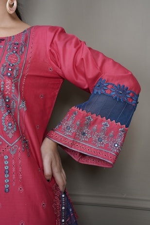 A detailed close-up of a garment with intricate embroidery. The fabric is a vibrant pink with elaborate patterns and embellishments highlighting the design. The sleeve features decorative blue lace with floral motifs, and the person is wearing large hoop earrings.