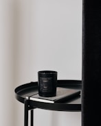 A black candle with a label sits on top of a closed book, both placed on a round black table. The table is next to a tall, dark curtain and a plain, light-colored wall.