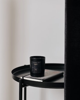 A black candle with a label sits on top of a closed book, both placed on a round black table. The table is next to a tall, dark curtain and a plain, light-colored wall.