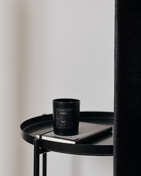 A black candle with a label sits on top of a closed book, both placed on a round black table. The table is next to a tall, dark curtain and a plain, light-colored wall.