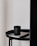 A black candle with a label sits on top of a closed book, both placed on a round black table. The table is next to a tall, dark curtain and a plain, light-colored wall.